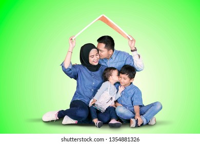 Muslim Family Holding A Roof Symbol From Cardboard Over Their Heads While Kissing Each Other In The Studio. Shot With Green Screen