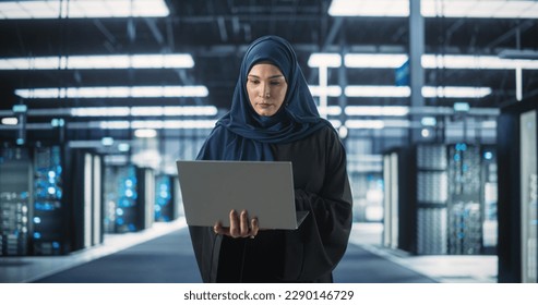 Muslim Female Data Center IT Engineer Standing in a Room with Server Racks. Cloud Computing Architect Uses Laptop Computer for Servicing the System in Cyber Security and Data Protection Facility
