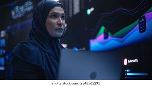 Muslim Female Data Center IT Engineer Standing in a Room with Statistics and Charts Settings on a Digital Screen. Cloud Computing Expert Uses Laptop for Servicing the System in Cyber Security Facility - Powered by Shutterstock