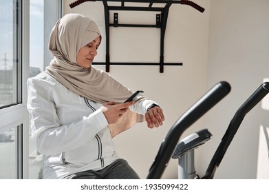 Muslim Fit Woman In Hijab Checking Her Fitness Track And Her Heart Rate After Cardio Workout On Stationary Bike.