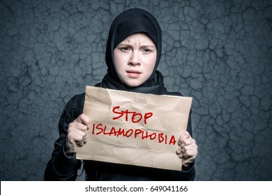 Muslim girl in black hijab holding a poster with an inscription STOP ISLAMOPHOBIA on the background wall with cracks