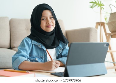 Muslim Girl Is Studying Online Via The Internet On Tablet While In The Living Room At Home Morning, Asian Elementary School Children Sitting Write Homework. Concept Distance Learning At Home