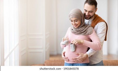 Muslim Husband Putting Tiny Shoes On Pregnant Wife's Belly Embracing Her Posing Standing At Home Near Window. New Life, Childbirth And Family Happiness. Panorama, Empty Space For Text - Powered by Shutterstock