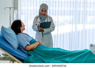 Muslim Islam Arab Young Professional Female Doctor Wearing Hijab And Stethoscope Holding Clipboard Writing Symptom Check Up Visiting Advising Senior Happy Patient Laying Down On Bed In Ward Room.