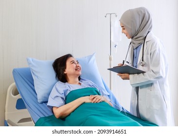 Muslim Islam Arab Young Professional Female Doctor Wearing Hijab And Stethoscope Holding Clipboard Writing Symptom Check Up Visiting Advising Senior Happy Patient Laying Down On Bed In Ward Room.