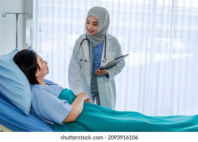 Muslim Islam Arab Young Professional Female Doctor Wearing Hijab And Stethoscope Holding Clipboard Writing Symptom Check Up Visiting Advising Senior Happy Patient Laying Down On Bed In Ward Room.