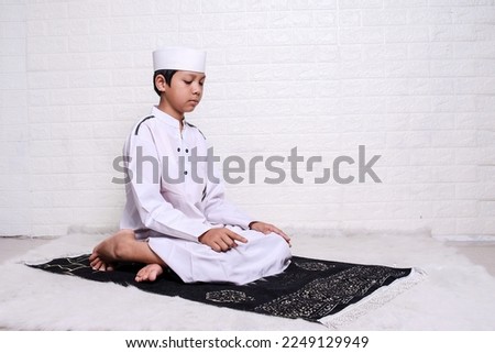 Muslim kid wearing white rob and skullcap praying salat in sitting position,  hold that the right index finger is raised when reciting the salawat. 
