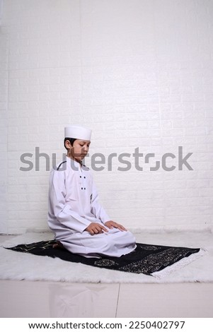 Muslim kid wearing white rob and skullcap praying salat in sitting position,  hold that the right index finger is raised when reciting the salawat. 