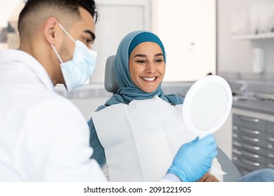 Muslim Lady In Hijab Looking At Her New Smile After Teeth Treatment In Dental Clinic, Middle Eastern Dentist Holding Mirror, Showing Result Of Medical Procedures For Islamic Female Patient, Closeup