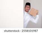 A muslim man peeking out behind wall carrying a cardboard box with excited expression