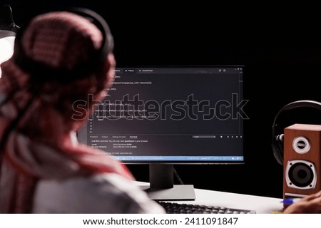 Muslim man sits at a computer and programs, exhibiting the app development process. Over-the-shoulder image of a computer monitor displaying code created by an Arab man dressed traditionally.