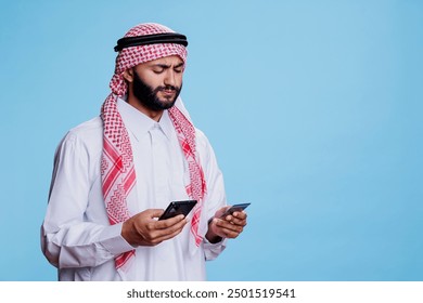 Muslim man in traditional clothes holding credit card and smartphone while shopping online. Arab with mobile phone making purchase in internet store using electronic banking - Powered by Shutterstock