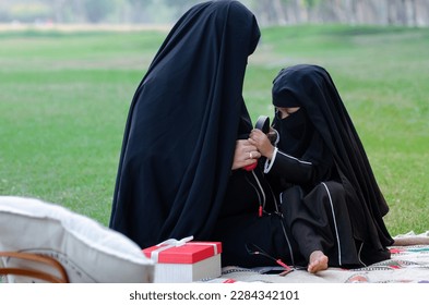 Muslim mother and daughter wearing black niqab spending weekends relaxing in the park, sitting on picnic blankets in the park, little girl using headphones