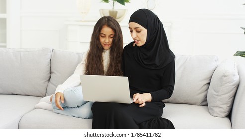 Muslim Mother In Headscarf Talking With Cute Teen Daughter, Sitting On Sofa And Using Laptop In Living Room. Pretty Arabian Woman In Hijab Watching Video On Computer And Browsing With Teenage Girl.
