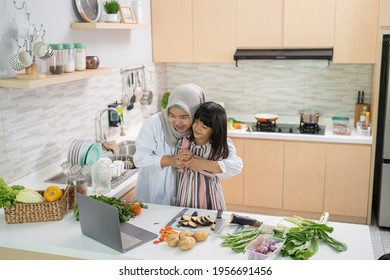 Muslim Mother Looking At Recipe From Laptop And Cooking With Her Daughter. Having Fun Woman With Hijab And Kid Preparing Dinner Together