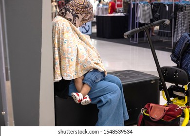 Muslim Mother Wearing Hijab Breastfeeding Her Child Under Cover Sitting In Public Area In Shopping Mall