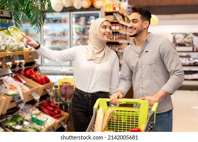 Muslim Spouses Doing Grocery Shopping Taking Vegetables From Shelf Buying Food In Supermarket. Wife Smiling To Husband Choosing Organic Products Standing With Shop Cart In Store. Consumerism