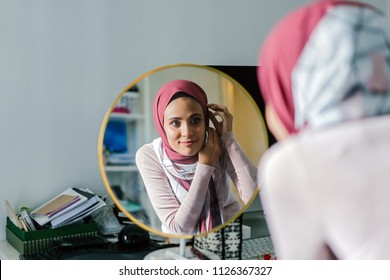 A Muslim woman adjusts her tudong (hijab head scarf) as she looks at herself into a round mirror in her home. Her headscarf is red orange and she is slim and attractive. - Powered by Shutterstock