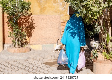 muslim woman with bags in hands in traditional long attire walks down street Red City Marrakech, authentic urban African landscape, daily activities people - Powered by Shutterstock