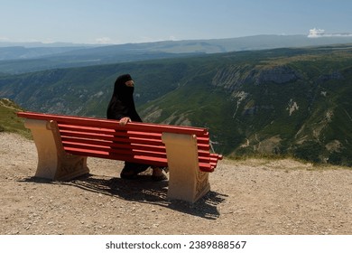 A Muslim woman in a black niqab sits on a bench and looks at the mountains from the back. Horizontal photo