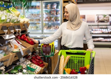 Muslim Woman Choosing Vegetables Doing Grocery Shopping In Supermarket, Wearing Hijab And Walking With Shop Cart Full Of Products. Commerce And Consumerism Concept