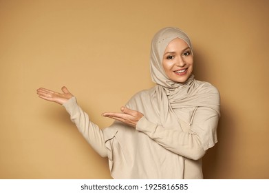 Muslim woman with covered head in hijab points side on beige background and smiles with toothy smile while looking at camera