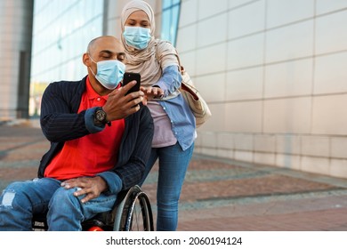 Muslim woman and disabled man wearing face mask using cellphone together outdoors
