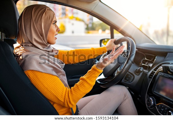 Muslim\
Woman is driving her car very aggressive and gives gesture with her\
hand. Closeup portrait displeased angry, aggressive Muslim woman\
driving car, shouting at someone, hands up in\
air
