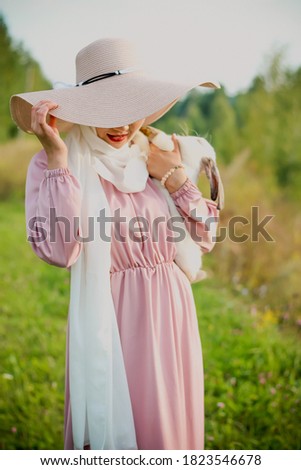 A Muslim woman in a hat is holding a white rabbit in her arms. A portrait of a Muslim woman in a pink dress and a scarf, a pearl bracelet. Tenderness. Soft focus.