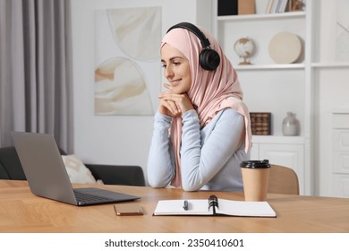 Muslim woman in hijab using laptop at wooden table in room - Powered by Shutterstock