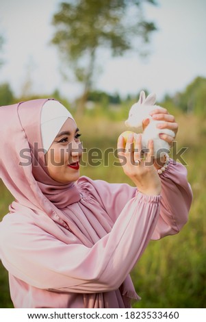 Muslim woman hold a small white rabbit in her arms. A portrait of a Muslim woman in a pink dress and a scarf, a pearl bracelet. Tenderness.