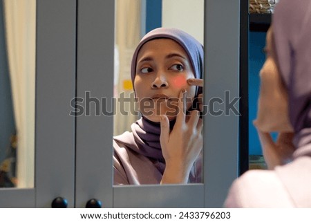 Muslim woman looking at herself in the mirror, holding her cheek due to acne