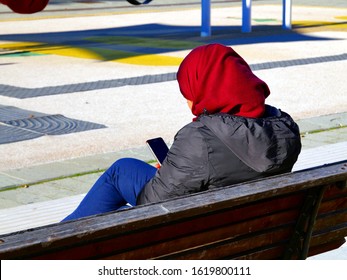 Muslim woman looks at cell phone sitting on a bench in the sun in the park photographed from behind - Powered by Shutterstock