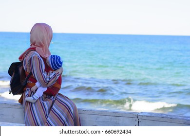 A Muslim Woman On The Beach With A Child. Asian Baby Carrier. 