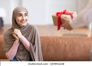 Muslim Woman Receiving Wrapped Gift Box From Man Sitting On Sofa At Home. Presents Delivery, Birthday And New Year Holiday Celebration Concept. Cropped, Selective Focus