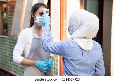 Muslim Woman Retail Business Staff Or Grocery Shop Keeper Checking Fever Of Asian Coworker By Portable Thermo Scan Device; Concept Of New Normal Business Practice