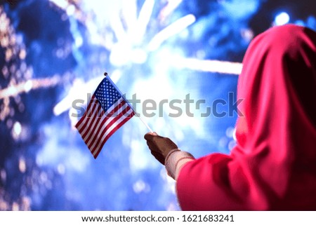 Muslim woman in a scarf holding American flag  during fireworks at night. 