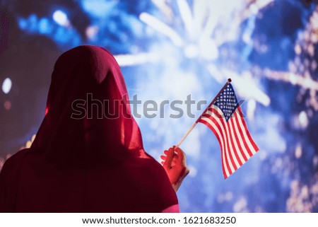 Muslim woman in a scarf holding American flag  during fireworks at night. 