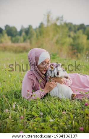 A Muslim woman is sitting on the grass and hold a two rabbit in her arms. A portrait of a Muslim woman in a pink dress and a scarf, a pearl bracelet. Tenderness. White and gray rabbit.