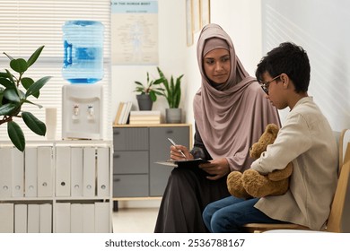 Muslim woman wearing hijab sitting with child holding teddy bear in office setting, engaging in supportive conversation. Scene includes water cooler and potted plants - Powered by Shutterstock