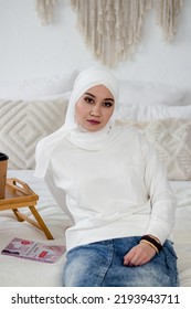 A Muslim Woman In A White Headscarf Sits On A Bed And In A Denim Skirt On The Bed Next To A Tray.