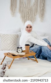 A Muslim Woman In A White Headscarf Sits On A Bed And In A Denim Skirt On The Bed Next To A Tray.