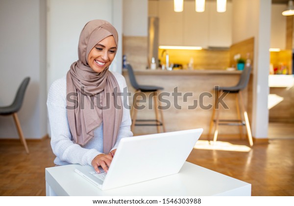 Muslim woman working with computer. Arab Young\
business woman sitting at her desk at home, working on a laptop\
computer and drinking coffee or tea. Muslim woman working at a home\
and using computer.