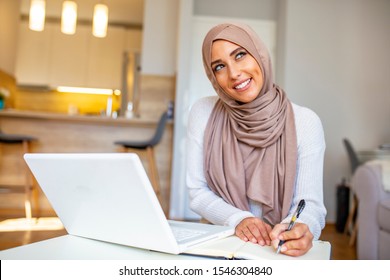 Muslim Woman Working In Home Office. Arabian Woman Dressed In Religious Veil Is Working On Her Laptop, Looking At Screen And Typing On Keyboard. She Seems To Be Concentrated And Glad 