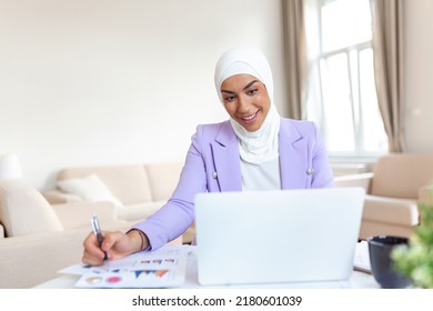 Muslim Woman Working In Home Office. Arabian Woman Dressed In Religious Veil Is Working On Her Laptop, Looking At Screen And Writing Notes.