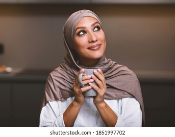 Muslim Woman's Dreams. Happy Arab Lady Drinking Coffee Dreaming And Thinking About Something Looking Aside Sitting In Modern Kitchen At Home, Wearing Hijab. Cozy Weekend Evening Concept