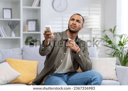 Muslim young man suffering from heat and discomfort at home, sitting on sofa holding remote control and turning on air conditioner.