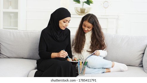 Muslim young mother in black hijab and abaya dress drawing and coloring picture with small teen daughter on couch in living room. Arabian woman and little girl painting together while playing at home. - Powered by Shutterstock