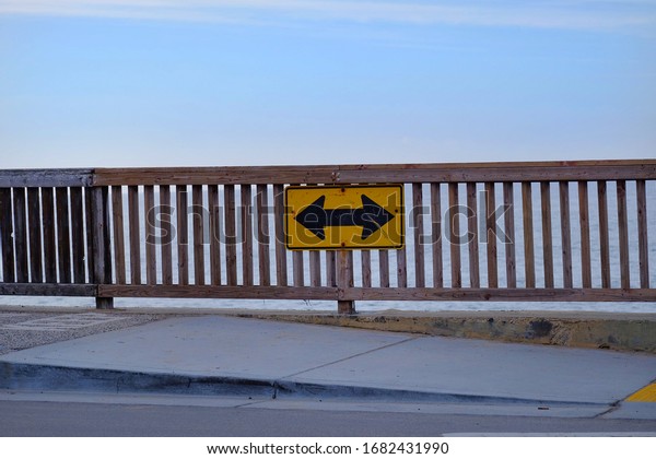 Must turn left or right\
directional road sign with opposing arrows and yellow background,\
with wooden fence along the coastline in the morning. Clear blue\
sky. 