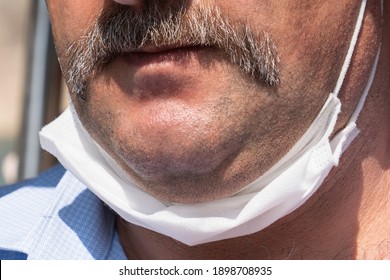 Mustached Man With A White Protective Medical Mask, Lowered To The Chin. Incorrect Wearing Of Medical Masks Under Quarantine. Closeup View.